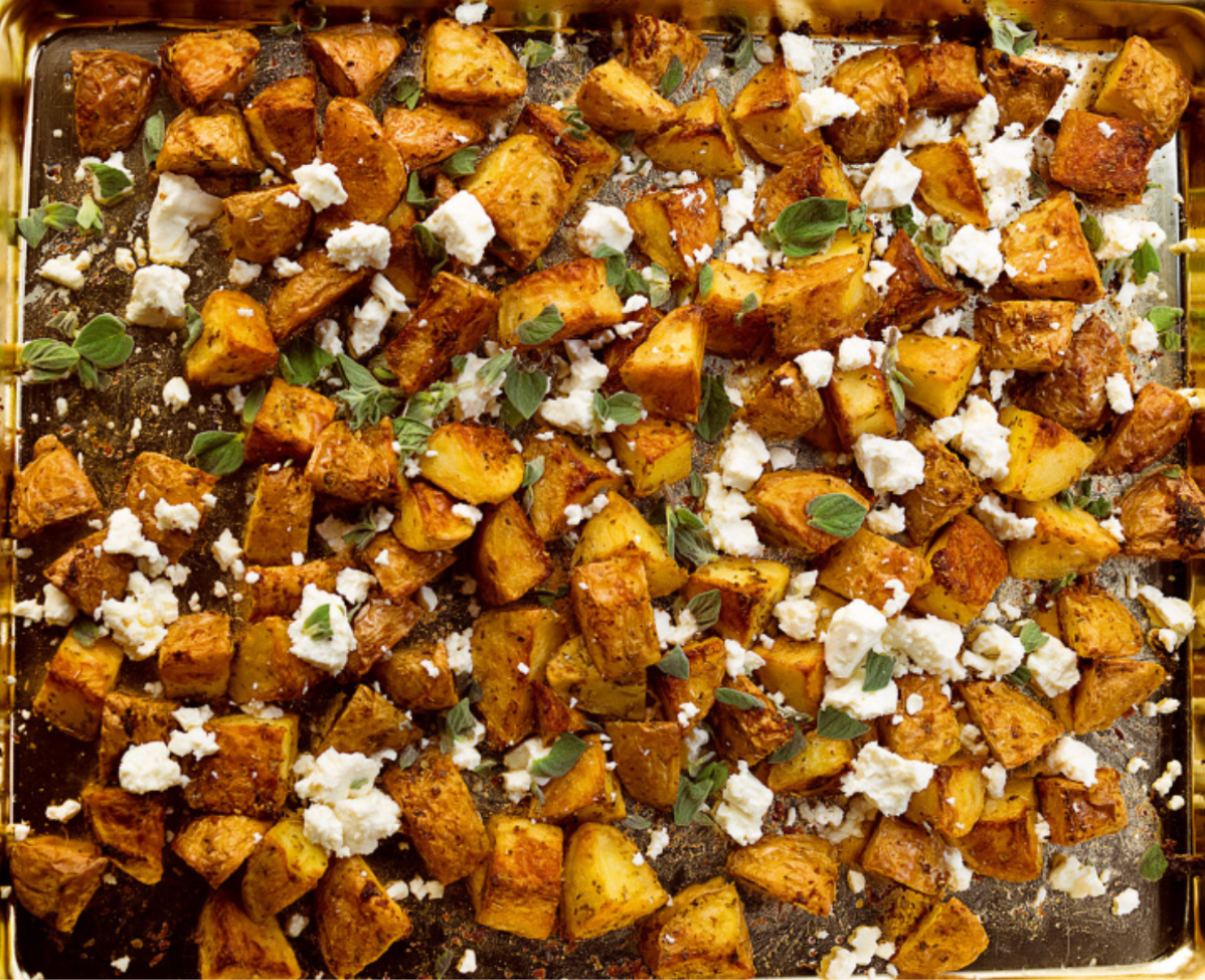 Overhead view of golden-brown roasted potato chunks spread on a metal baking tray. Crumbled white cheese and small green herb leaves are scattered over the potatoes.
