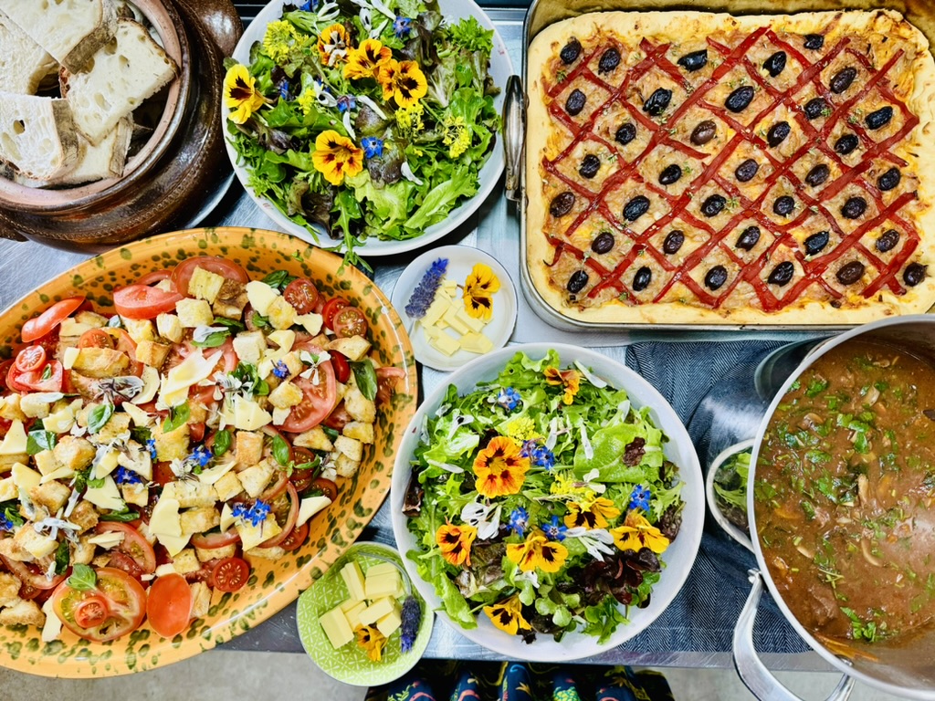 Overhead view of a table set with bowls of leafy salads topped with edible flowers, a tomato-and-crouton salad, sliced bread, small dishes of cheese, a baked tart with a red lattice and olives, and a pot of soup or stew.