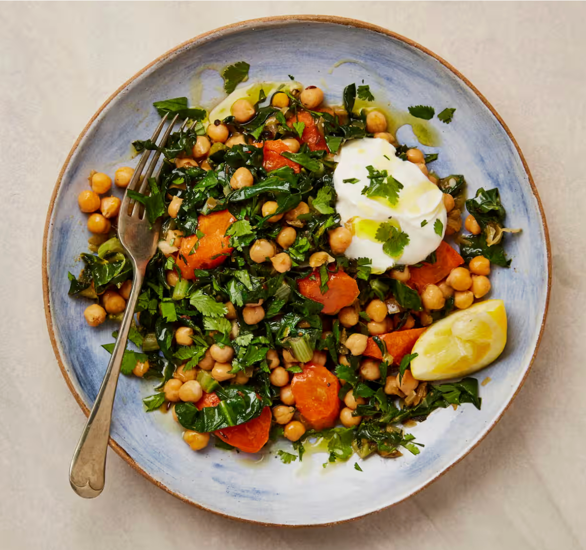 Overhead view of a blue bowl filled with a chickpea and leafy greens salad with orange vegetable slices and chopped herbs. A dollop of yogurt, a lemon wedge, and a fork rest in the bowl.