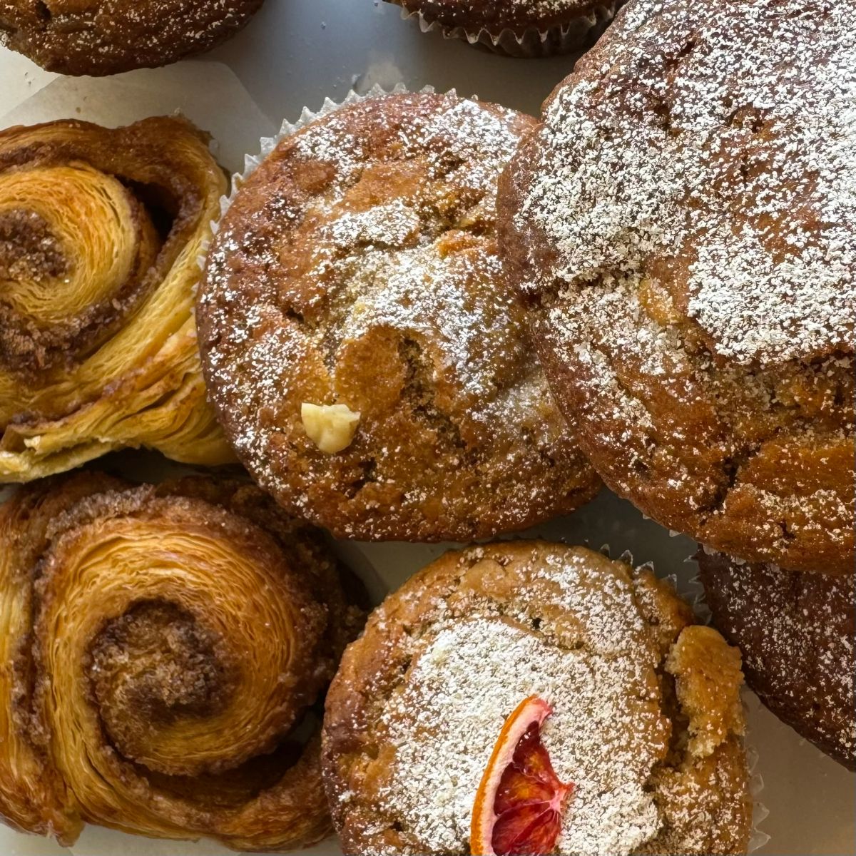 Close-up of assorted baked pastries in a box, including spiral Danish pastries and large muffins or cakes dusted with powdered sugar. One muffin is topped with a slice of dried citrus.
