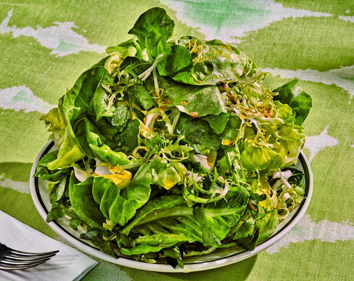 A heaped bowl of mixed green salad leaves with sprouts and thin slices of onion, drizzled with dressing. The bowl sits on a green patterned tablecloth with a fork and folded napkin beside it.