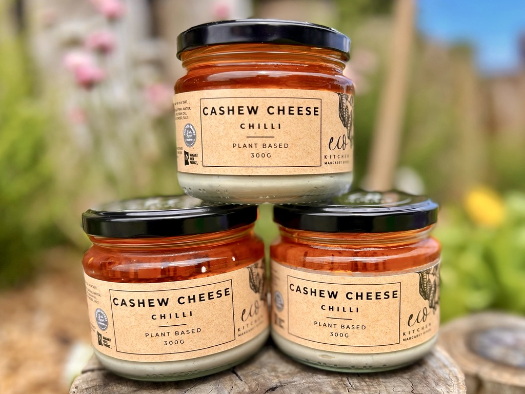 Three glass jars of “Cashew Cheese Chilli” are stacked in a pyramid on a wooden stump, each with a black lid and beige label. The background is an out-of-focus garden scene with greenery and flowers.