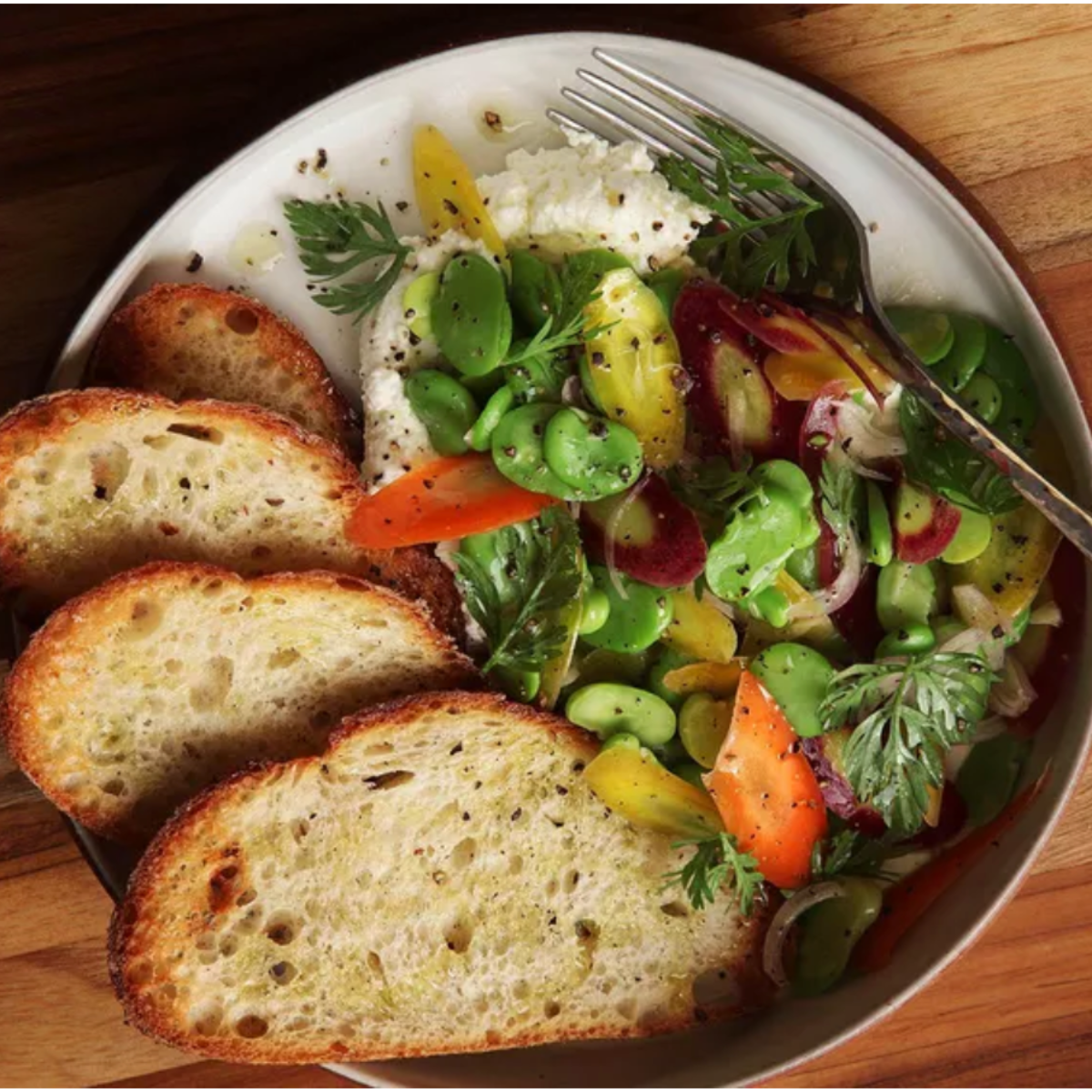 Overhead view of a bowl with a colorful salad of sliced vegetables, herbs, and a dollop of soft white cheese, sprinkled with black pepper. Several slices of toasted bread line one side, with a fork resting on the rim.