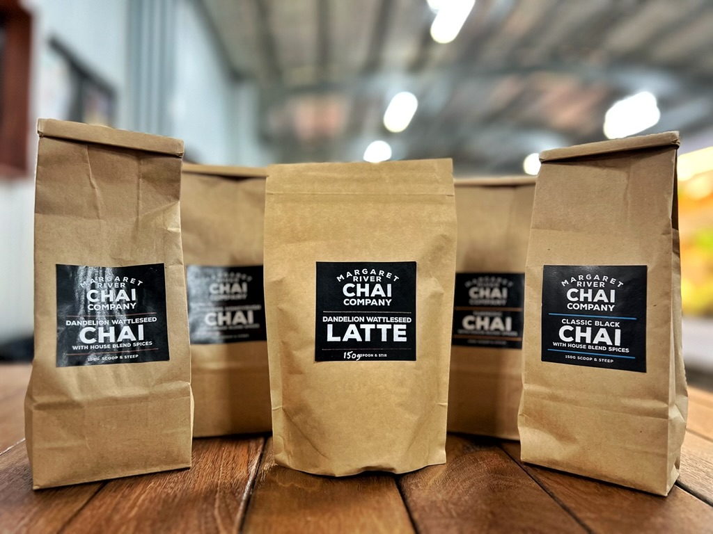 Brown paper bags of Margaret River Chai Company products on a wooden table, with labels for “Dandelion Wattleseed Latte” and “Chai” visible on the front bags. More similar bags are arranged behind them in a blurred indoor setting.