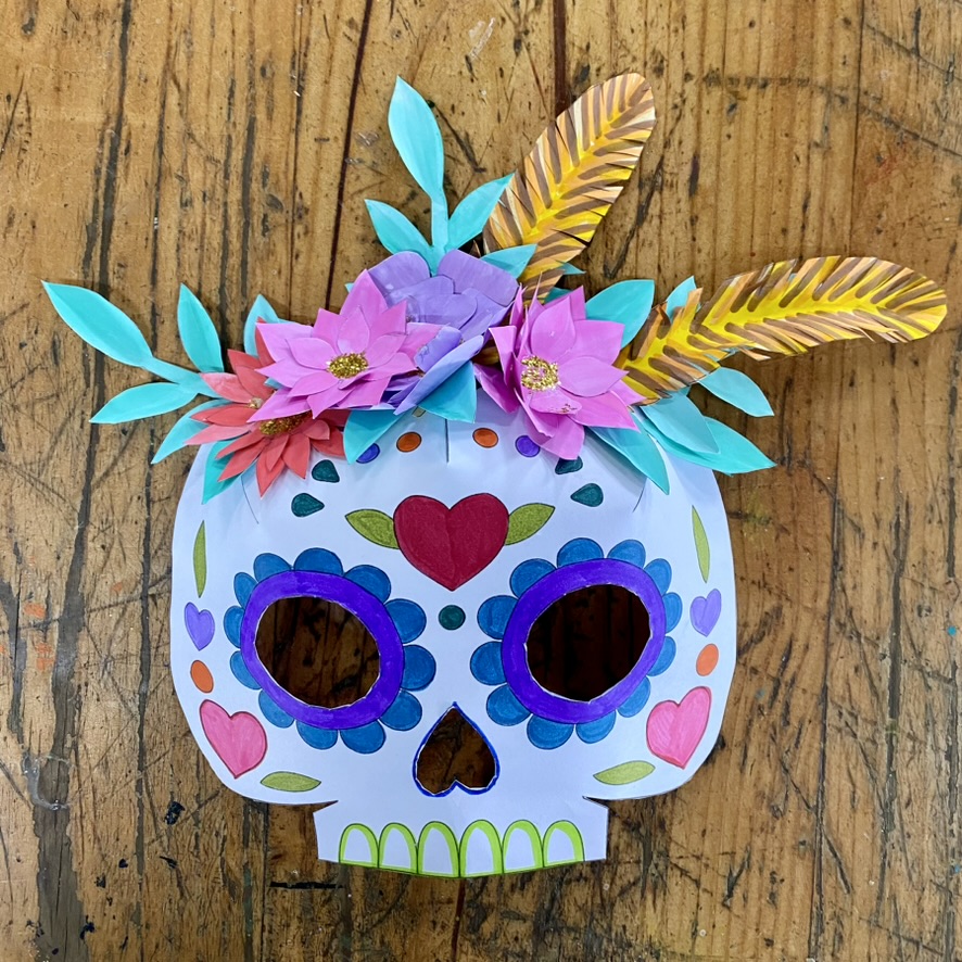 A colorful paper skull mask with purple eye cutouts, heart and dot decorations, and a crown of paper flowers and teal leaves. The mask sits on a scratched wooden tabletop.
