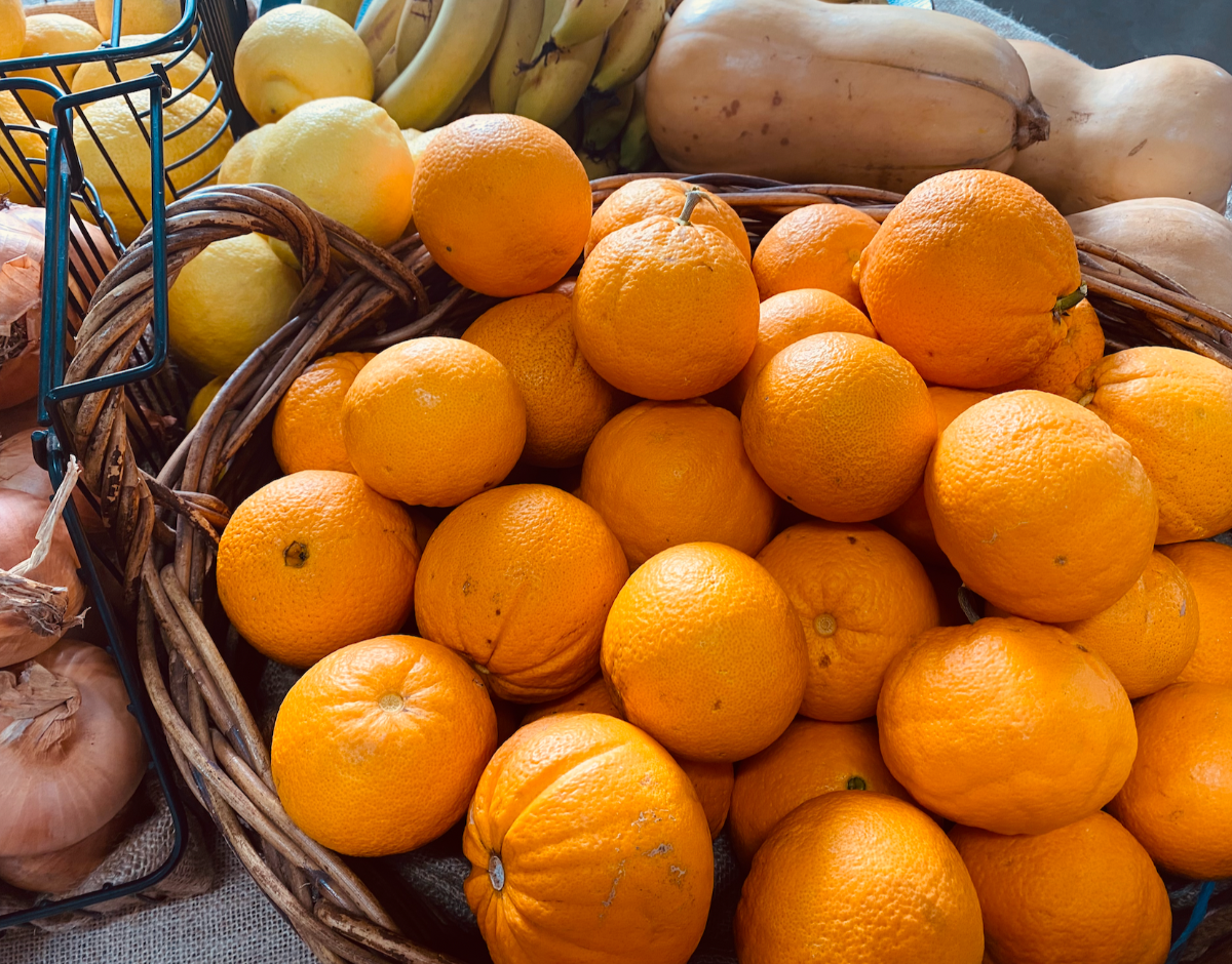 Wicker baskets filled with oranges and lemons, with loose onions at the left edge. Bananas and pale butternut squash are stacked in the background.
