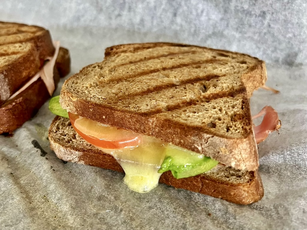 Close-up of a grilled sandwich on dark bread with melted cheese, tomato, lettuce, and a slice of cured meat on parchment paper. A second sandwich is partially visible in the background.