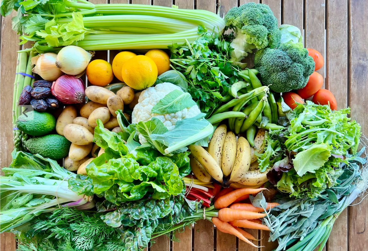 A wooden crate filled with produce including celery, broccoli, cauliflower, tomatoes, carrots, potatoes, onions, oranges, bananas, avocados, salad greens, herbs, and green beans. The crate sits on a wooden slatted surface.
