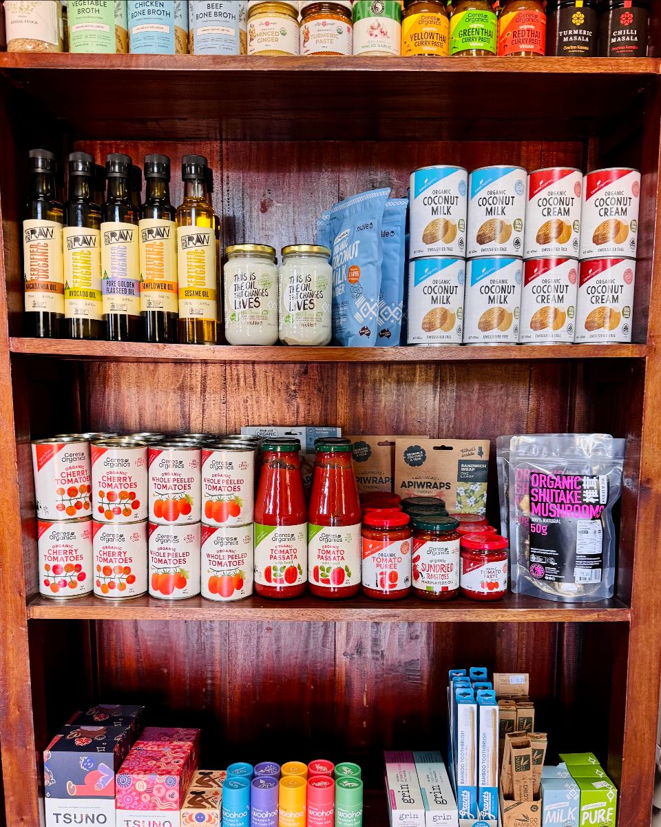 Wooden shop shelves stocked with pantry items, including bottled cooking oils, jars, canned tomatoes, and boxed coconut milk and coconut cream. The display also includes tomato sauces, wraps, and a bag of organic shiitake mushrooms.