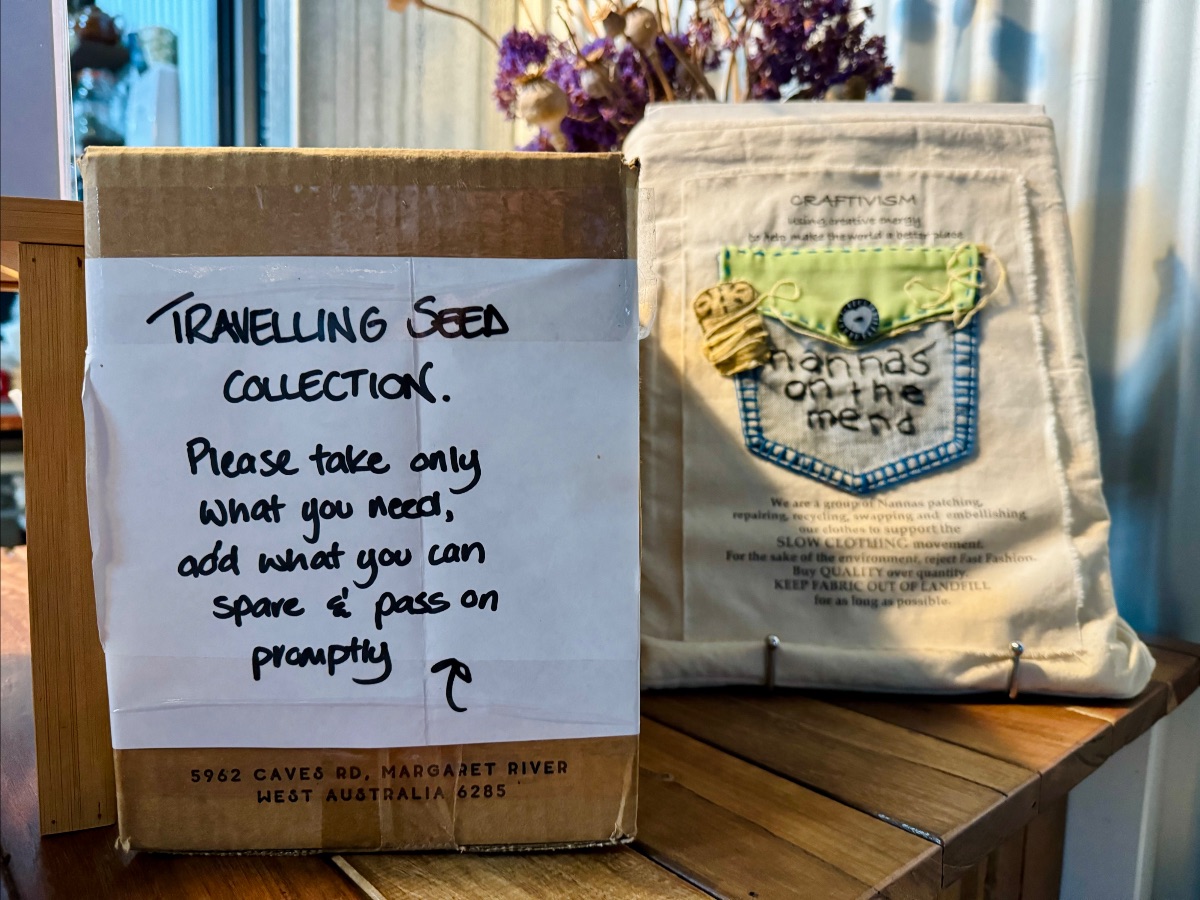 Cardboard box with a handwritten sign reading “Travelling Seed Collection” and instructions to take only what you need and add what you can spare. Beside it sits a fabric bag labeled “Craftivism” with a stitched patch reading “nannas on the mend,” on a wooden counter with dried flowers in the background.