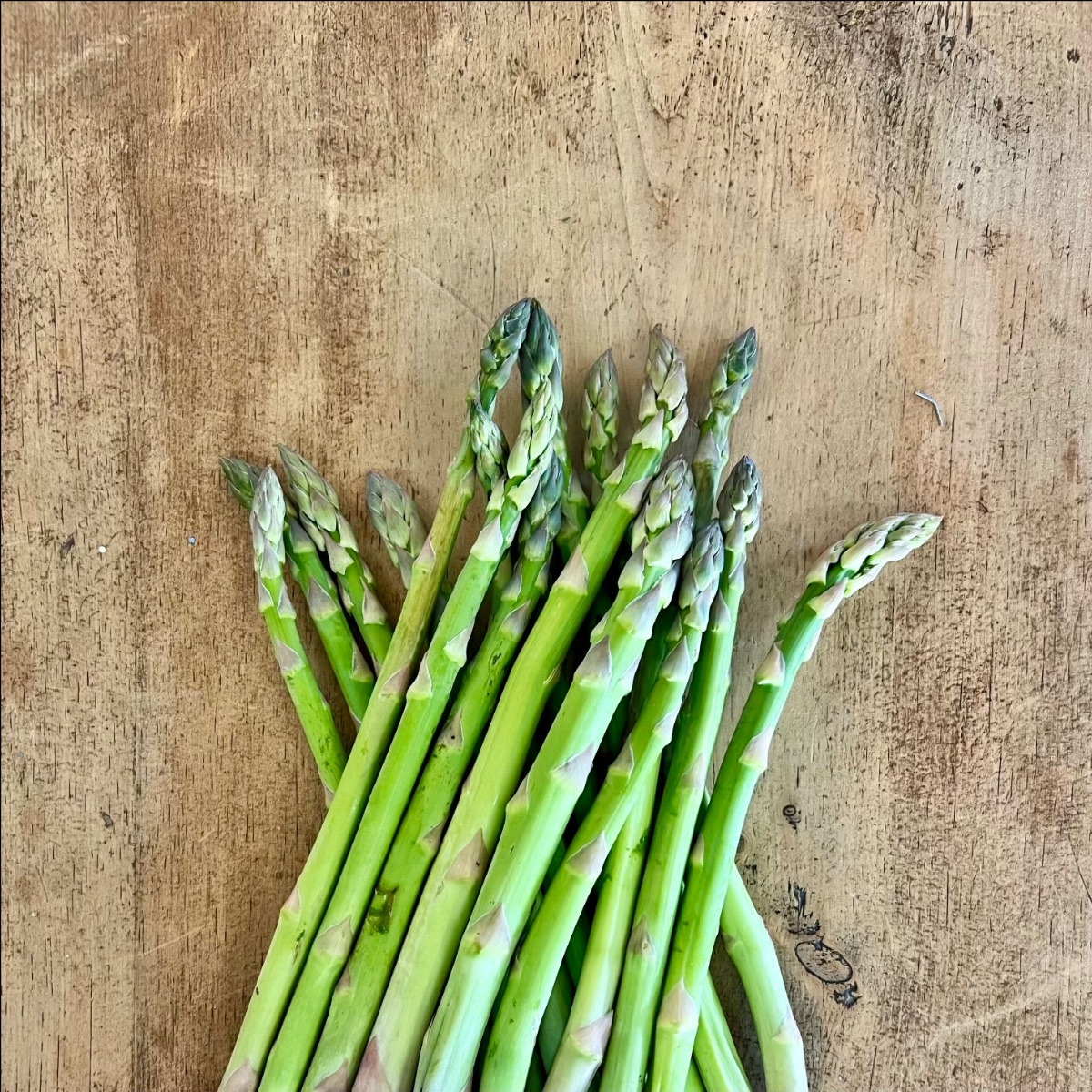 A small bunch of green asparagus spears lies on a worn wooden surface. The tips point upward and the stalks overlap in the center.