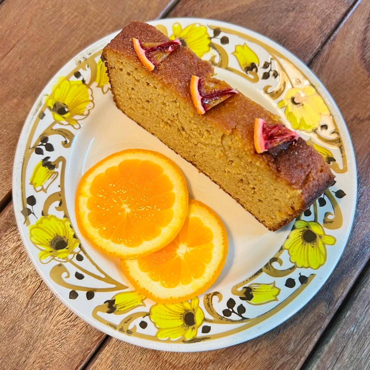 Slice of loaf cake topped with small citrus pieces on a floral-patterned plate. Two round orange slices sit beside the cake on a wooden table.