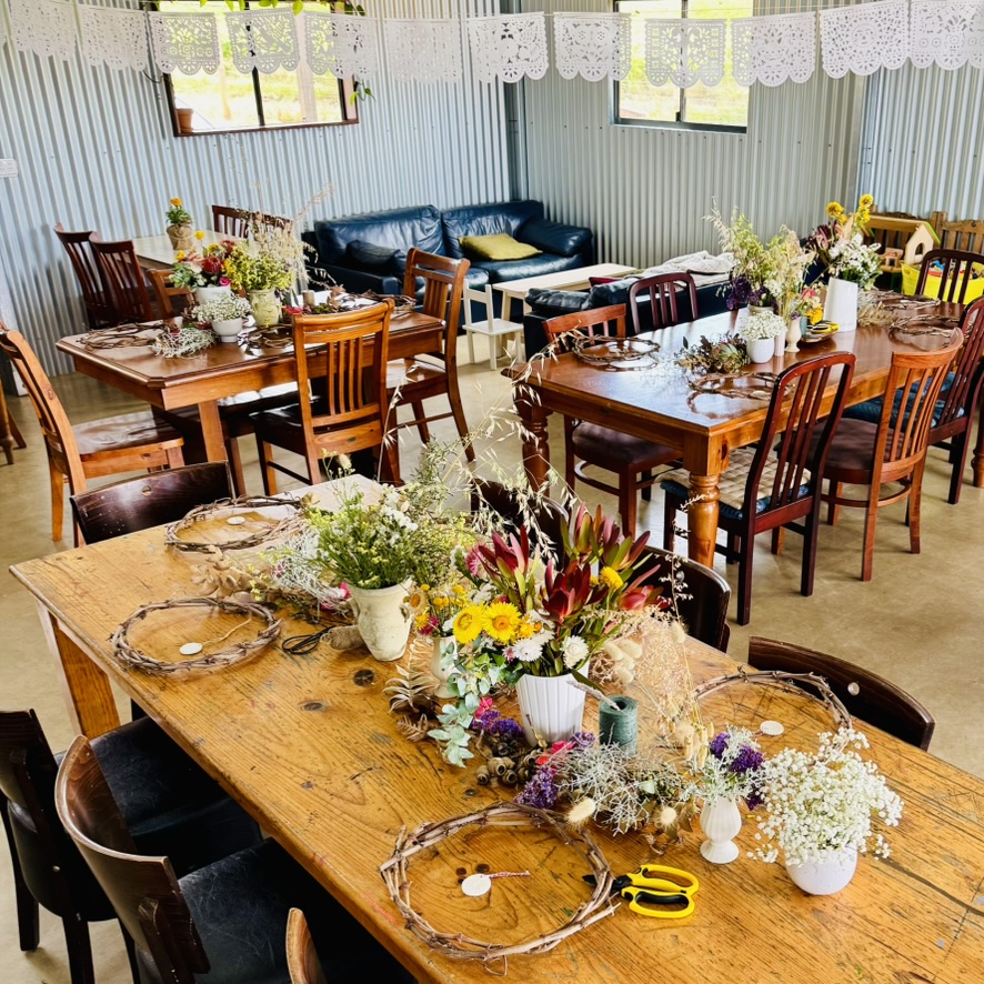 Several wooden dining tables with chairs are set up indoors, each decorated with floral arrangements and twig wreaths. A dark couch sits against a corrugated metal wall under windows, with white paper bunting hanging across the room.