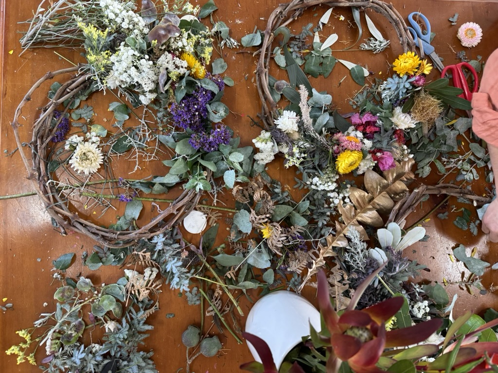 Overhead view of a wooden table covered with greenery and assorted flowers, with two grapevine wreath rings partially decorated. A pair of red scissors and blue snips sit on the right, and hands in a pink sleeve work near the wreath.