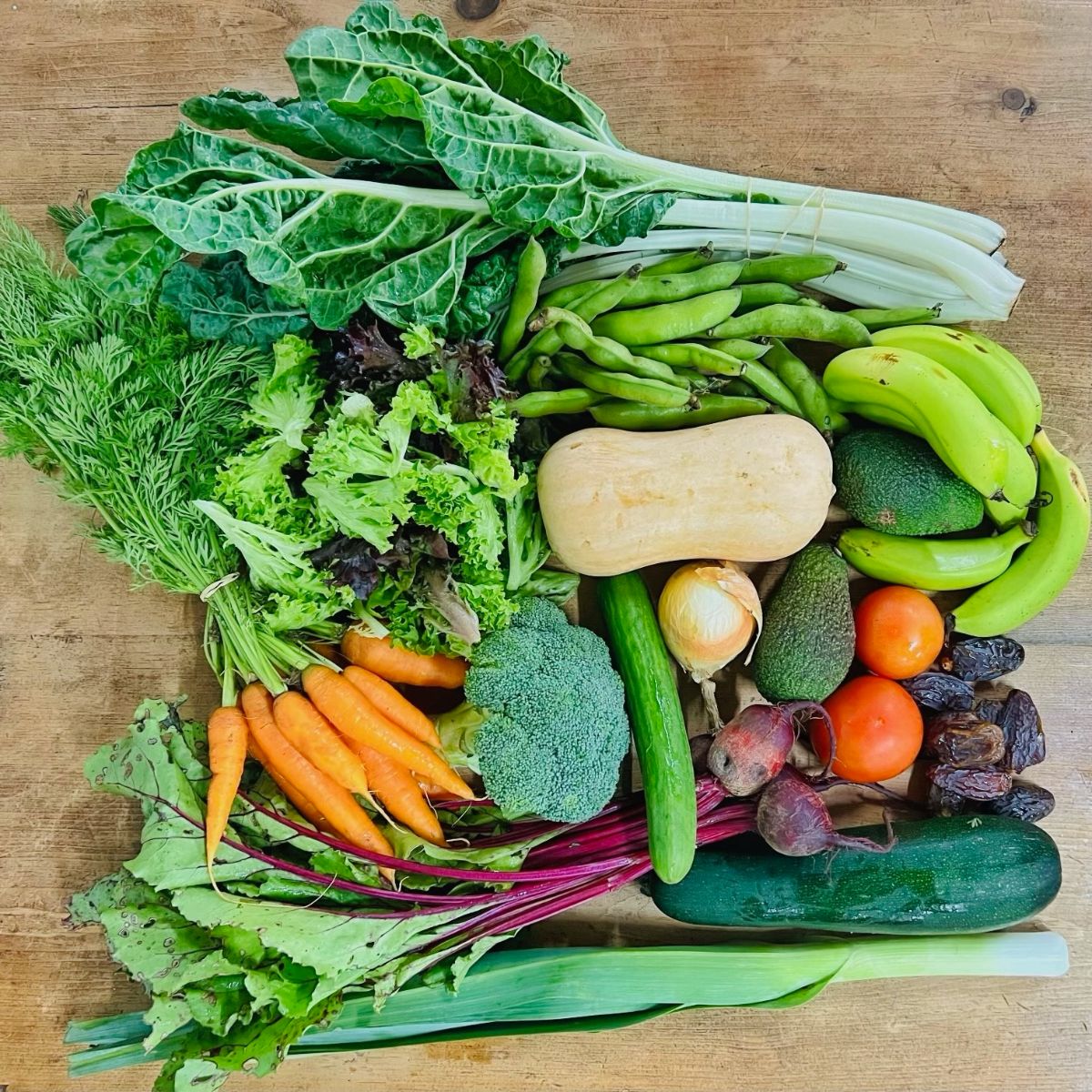 Assorted produce on a wooden table, including leafy greens, carrots, broccoli, beets, green beans, cucumbers, a dark green zucchini, and a butternut squash. Also shown are bananas, avocados, tomatoes, dates, an onion, and leeks/swiss chard stems.