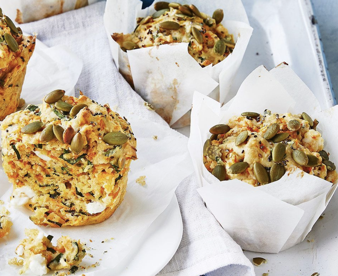 Three savory muffins in white parchment wrappers sit on a white tray and cloth napkin, topped with pumpkin seeds and small dark seeds. One muffin is cut open, showing green zucchini strands and white cheese pieces inside.
