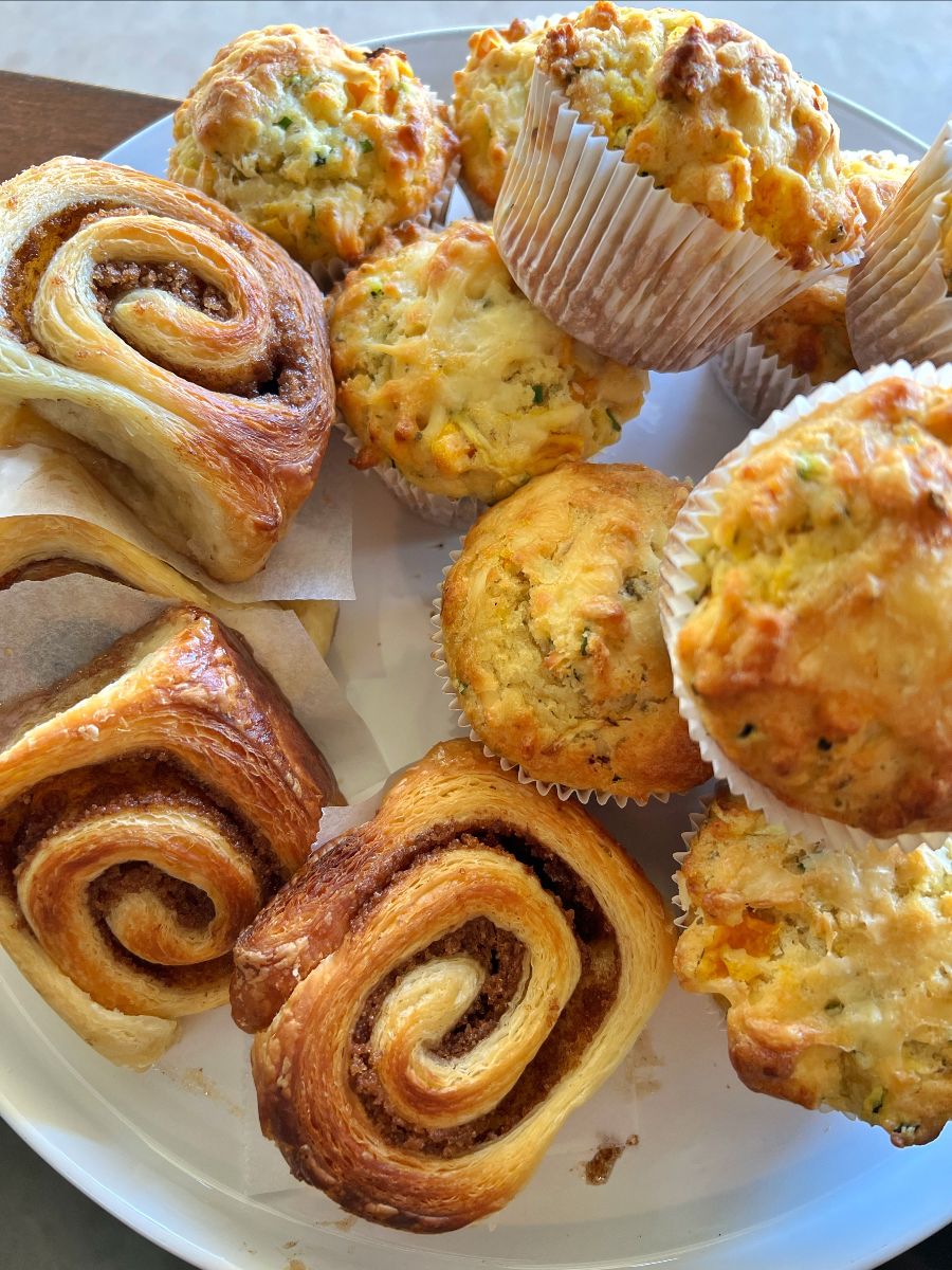 Close-up of a white plate holding several spiral pastries and a selection of golden muffins in paper cases. The baked goods fill most of the frame, showing flaky layers and browned tops.
