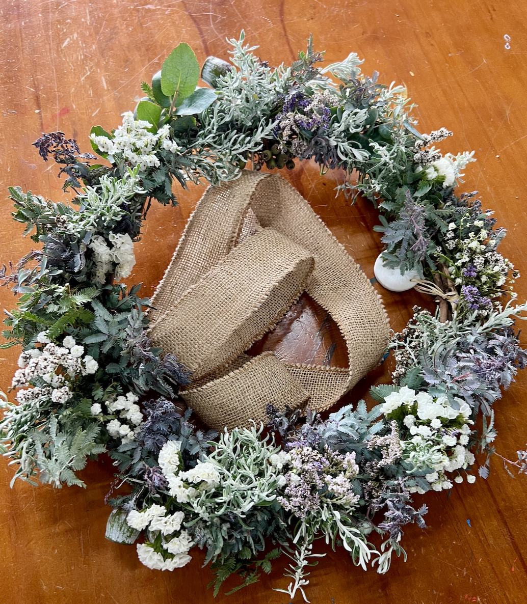 A circular wreath made of mixed greenery and small white and purple flowers sits on a wooden table. A wide burlap ribbon is draped through the center of the wreath.