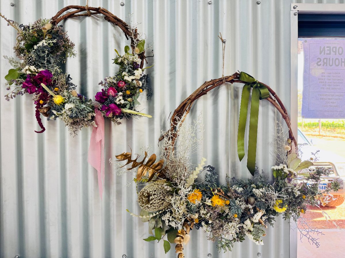 Two floral wreaths hang on a corrugated metal wall: one with colorful flowers and a pink ribbon, and a larger one with dried greenery, yellow blooms, and a green ribbon. A window at right shows part of an “OPEN HOURS” sign.