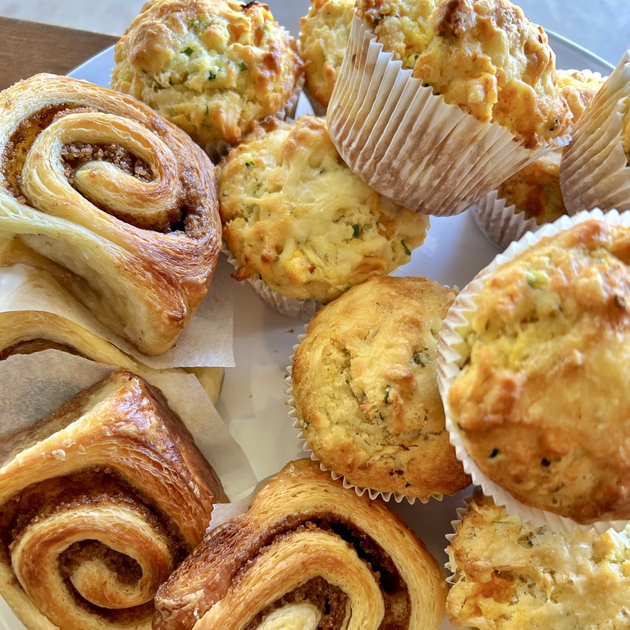 Close-up of a white plate filled with golden baked pastries: spiral cinnamon-style rolls and savory muffins in white paper liners. The muffins show melted cheese and small green and orange bits on top.