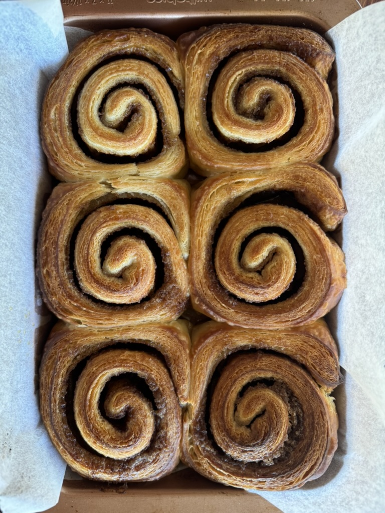 Top-down view of six golden-brown spiral pastries arranged in two columns inside a rectangular cardboard tray lined with white parchment paper. The pastries have visible layered swirls and browned edges.