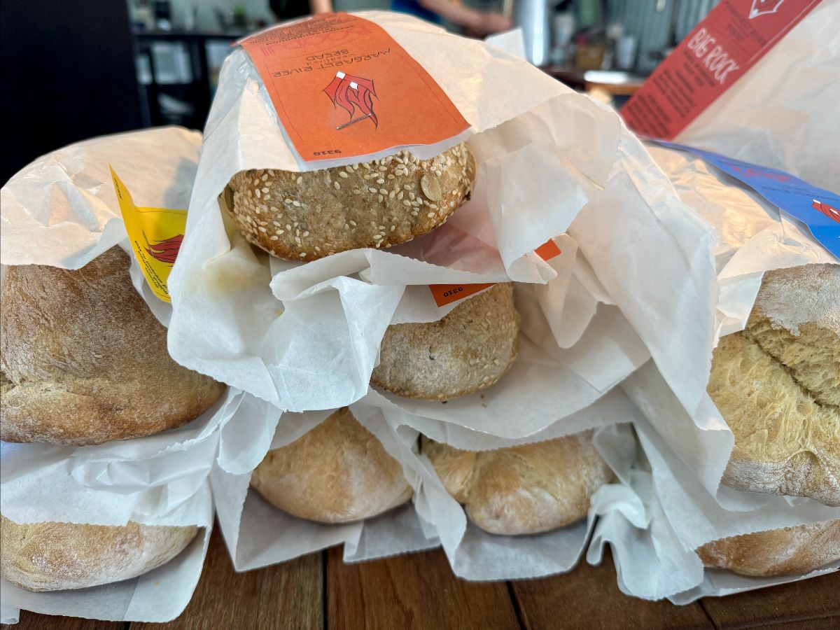 Several crusty bread rolls sit on a wooden table, each partly wrapped in white paper bags with colorful labels. One roll is topped with sesame seeds, and the background is softly blurred.
