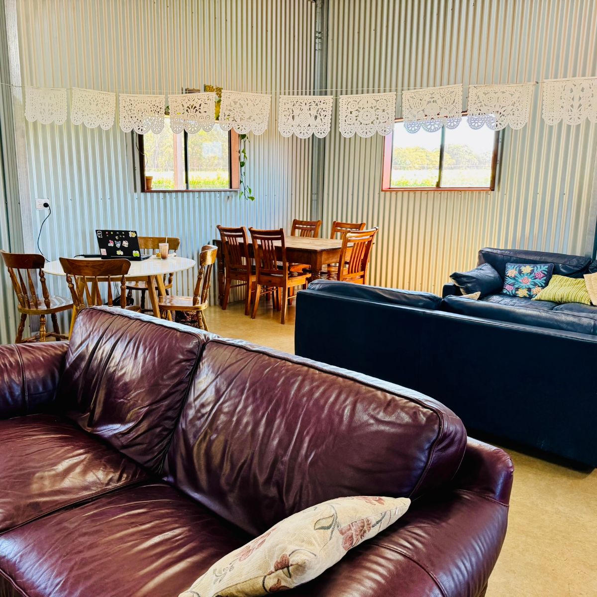 Indoor seating area with corrugated metal walls, two leather couches, and wooden tables and chairs. A round table holds an open laptop and a drink, with white paper cutout bunting hanging across the room and two windows in the back wall.