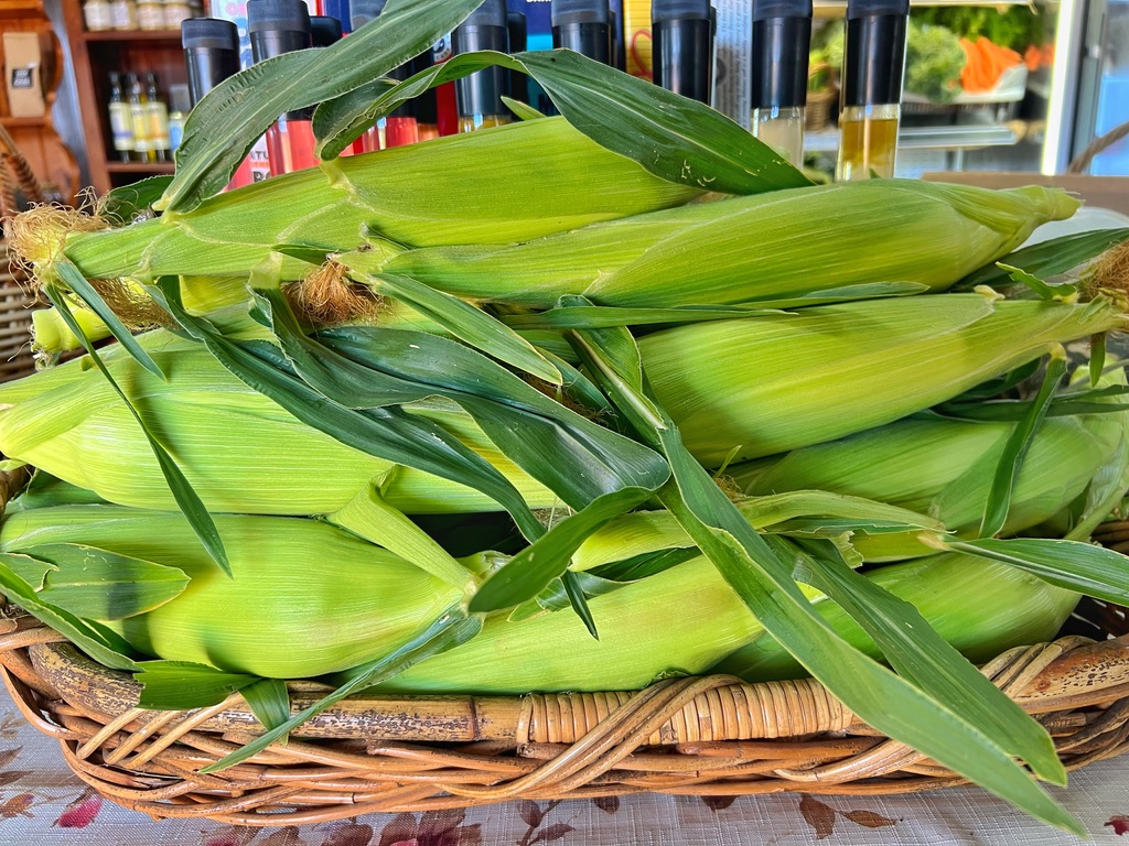 A wicker basket piled with fresh ears of sweetcorn in green husks, with some corn silk visible. Bottles and shelves are blurred in the background of the shop.