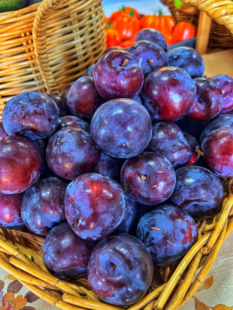 Close-up of a wicker basket filled with dark purple plums on a patterned tablecloth. Another basket is visible behind it with orange-red tomatoes in the background.