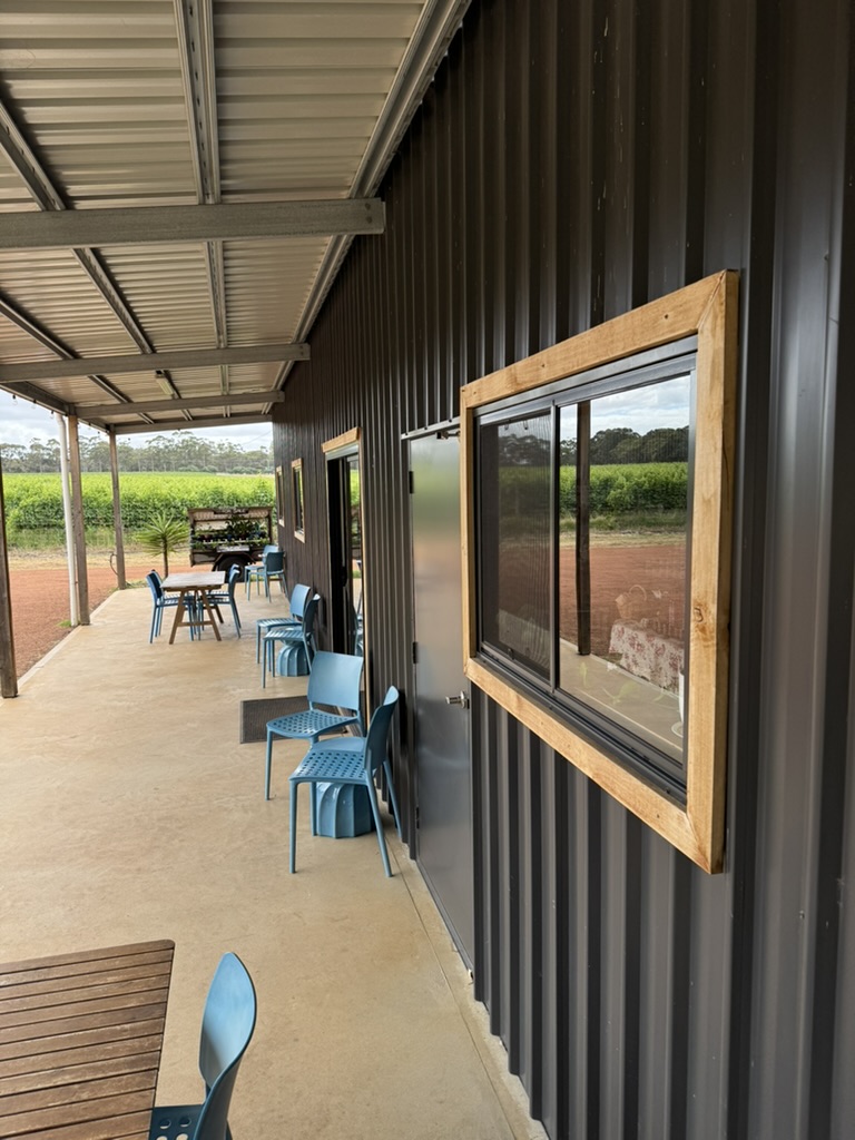 Covered outdoor walkway beside a black corrugated-metal building, with blue chairs and wooden tables lined up along the wall. Windows with wood trim reflect fields and trees in the background.
