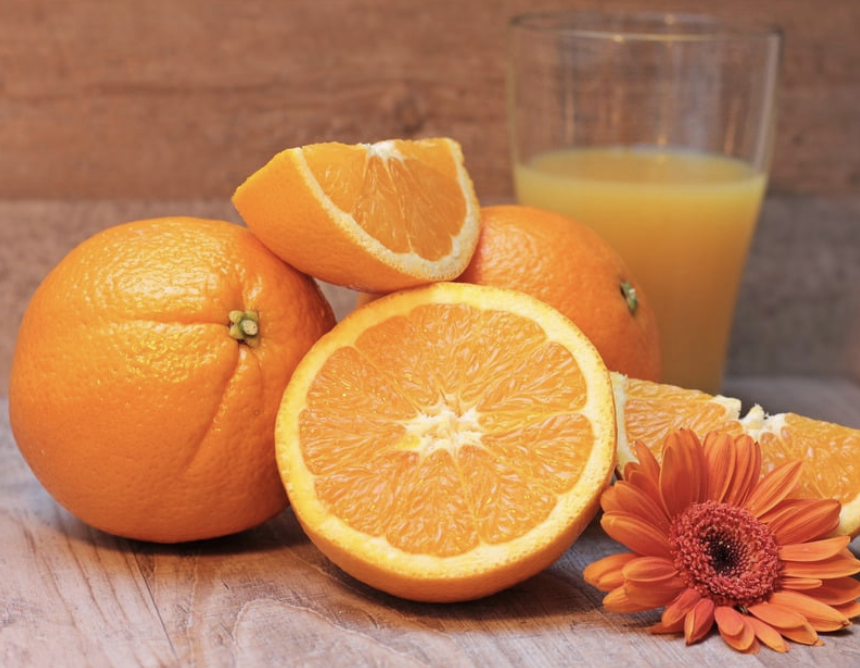 Whole and sliced oranges on a wooden surface, with an orange wedge on top and a halved orange in front. A clear glass of orange juice sits in the background, and an orange flower lies beside the fruit.