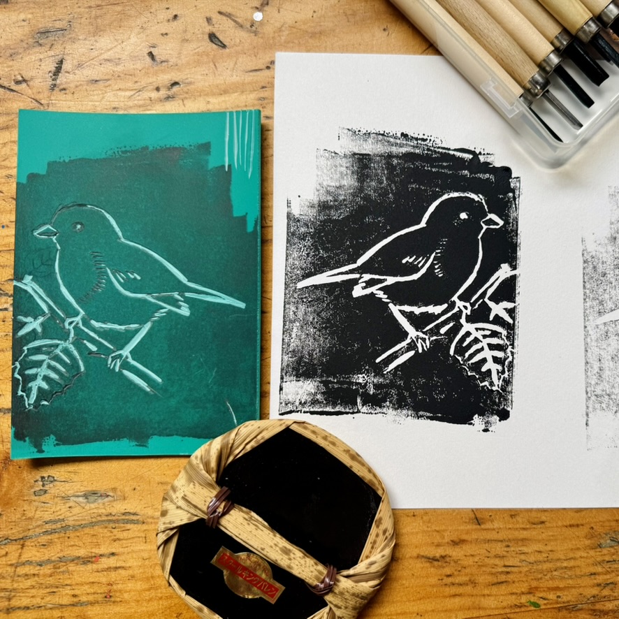 Green printing plate and a black-and-white bird print on paper sit on a wooden table. Carving tools and a small ink tray are placed nearby.