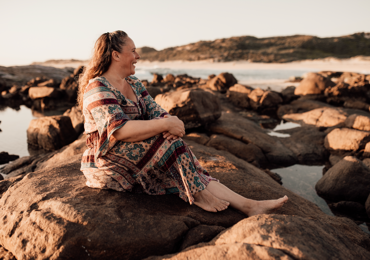 A woman in a patterned dress sits barefoot on large rocks by the ocean, looking to the side and smiling. Tide pools and a sandy shoreline are visible in the background.