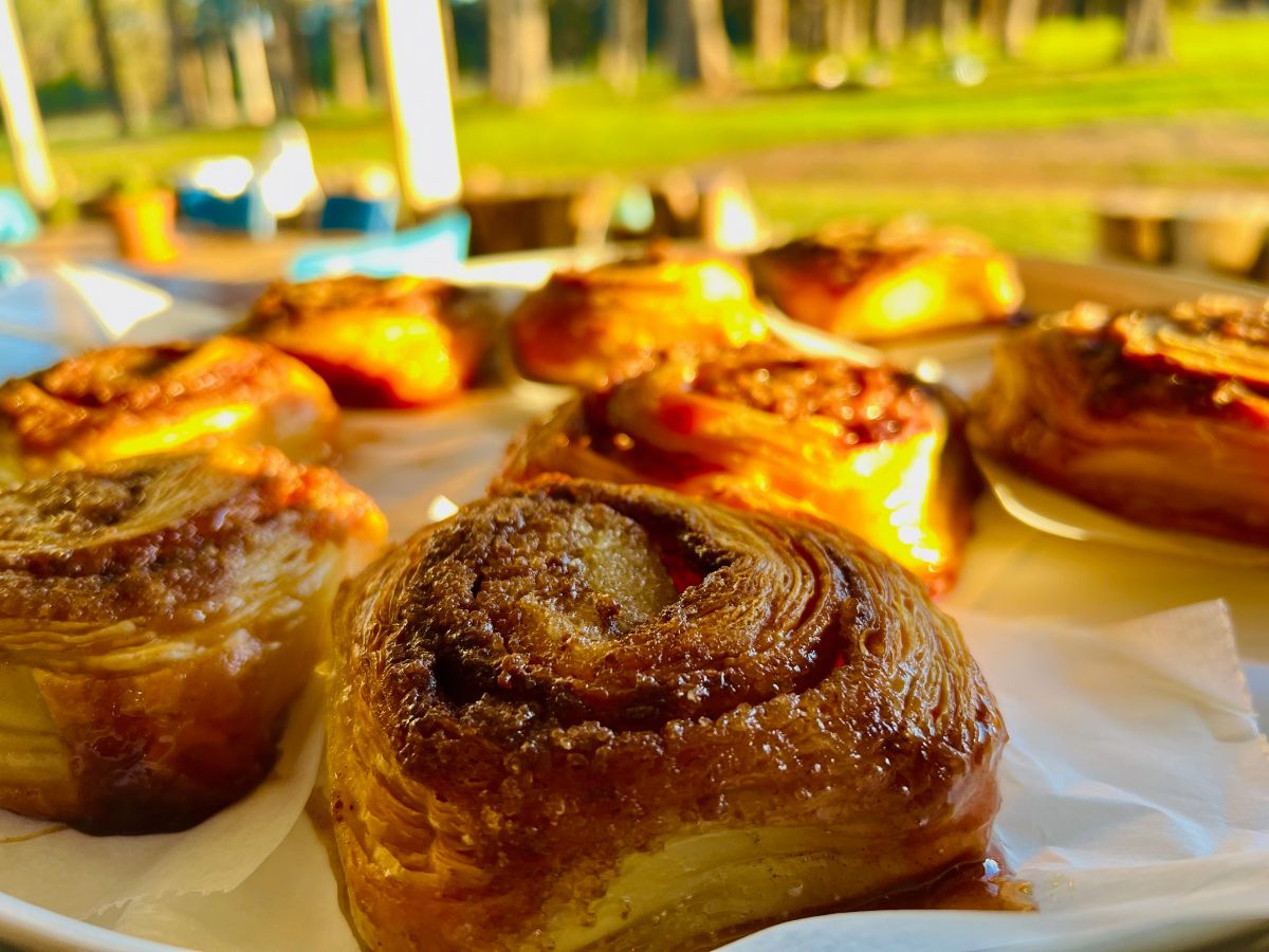 Close-up of several golden-brown spiral pastries on parchment paper on a table. The background is blurred, showing green grass and trees in sunlight.