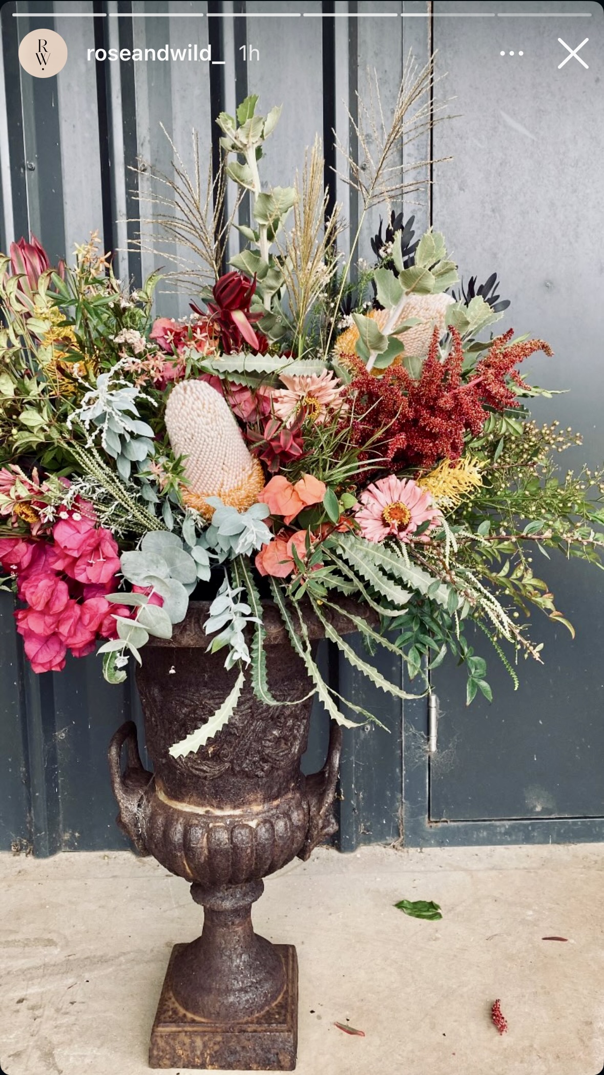 Large mixed flower arrangement with pink, red, and orange blooms and leafy greenery displayed in a dark metal urn. The urn sits on a concrete floor in front of tall gray metal doors.