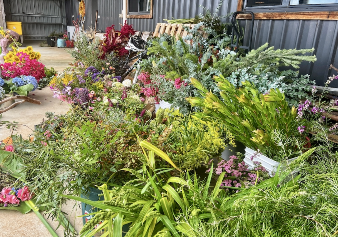 Large piles of assorted cut flowers and greenery in buckets and loose on the floor inside a corrugated-metal shed. A small child sits at the left beside bright bouquets, with pallets and a hand truck in the background.