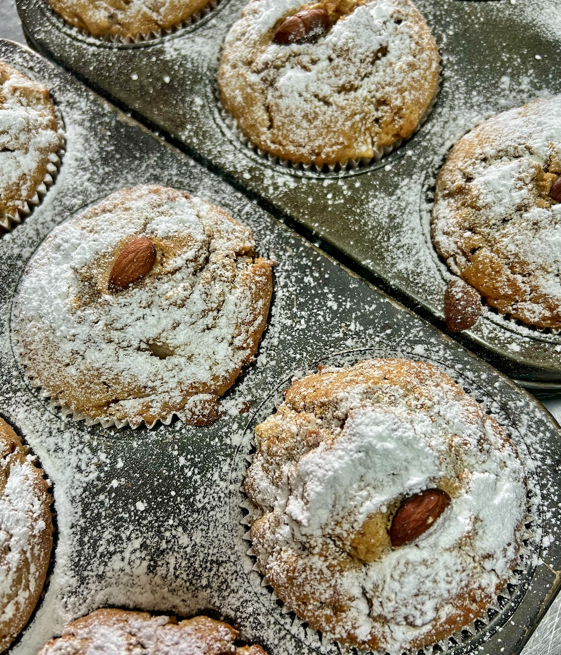 Close-up of several muffins in metal baking trays, heavily dusted with powdered sugar. Each muffin has an almond on top, with sugar scattered across the pans.