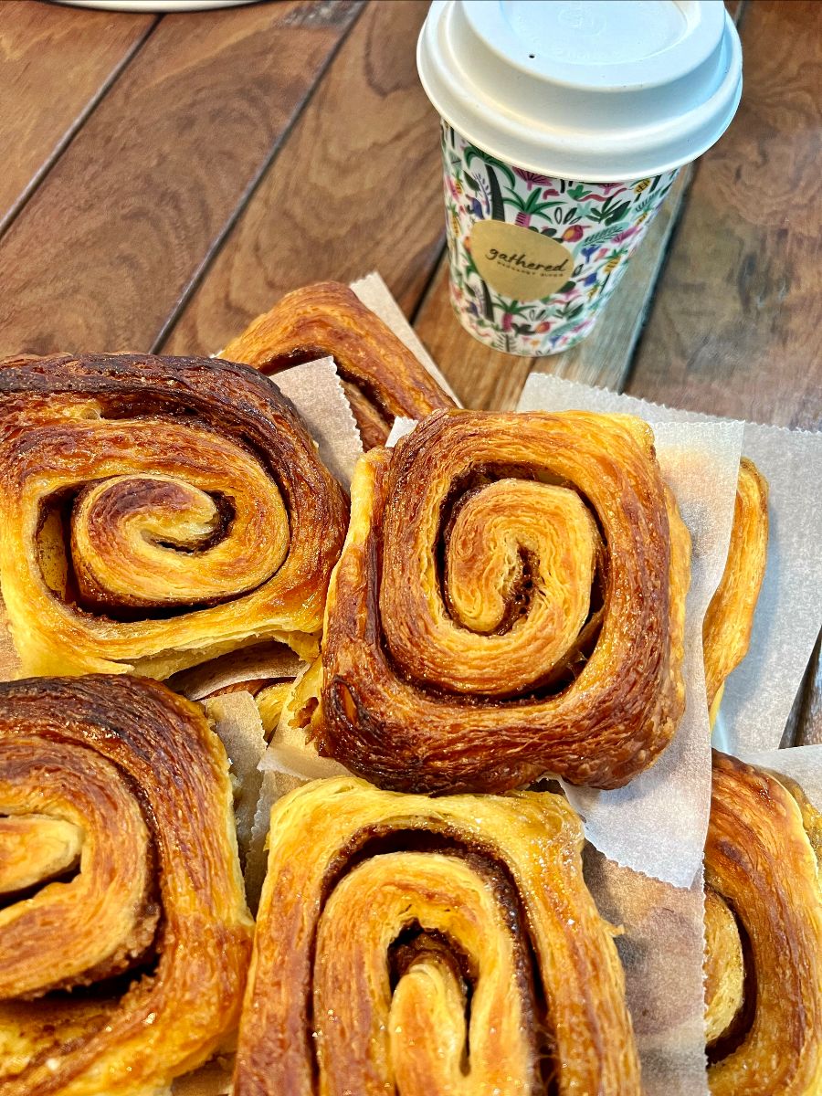 Close-up of several spiral-shaped, golden-brown pastries on parchment paper on a wooden table. A floral-patterned takeaway coffee cup with a white lid sits behind them.