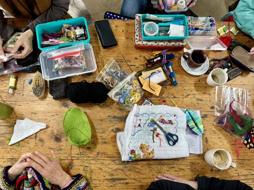 Overhead view of a wooden table covered with craft supplies, including embroidery floss in plastic boxes, scissors, needles, and a partially stitched fabric piece. Several people’s hands are visible around the table, along with balls of yarn and cups of coffee/tea.