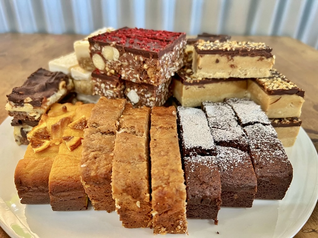 Assorted homemade traybakes and slices stacked on a white plate, including chocolate-topped bars, nut-studded squares, and brownies dusted with powdered sugar. The plate sits on a wooden table with a softly blurred background.