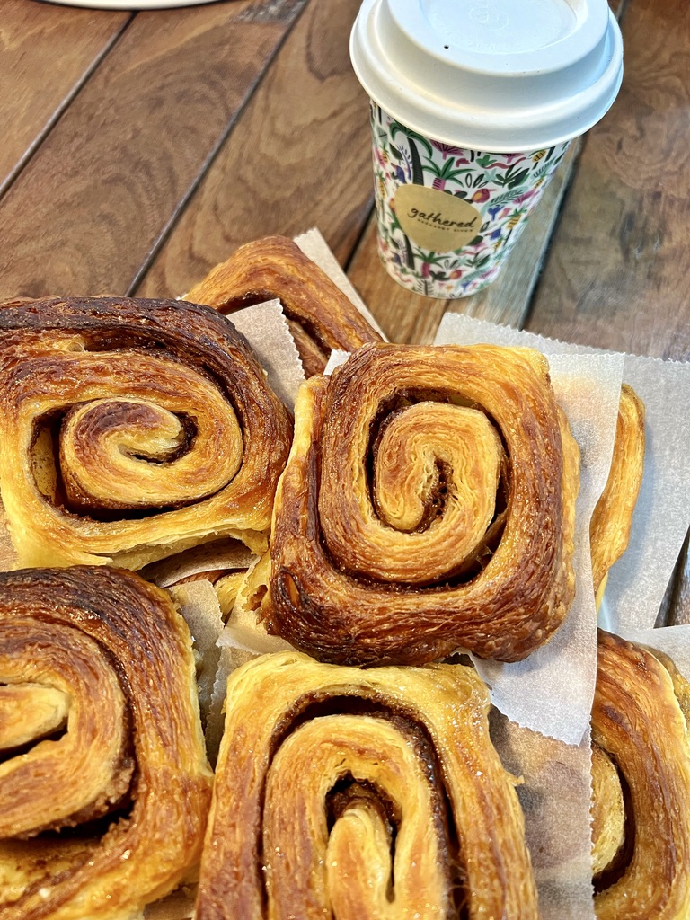 Close-up of several golden-brown swirled pastries on parchment paper on a wooden table. A floral-patterned takeaway coffee cup with a white lid sits in the background.