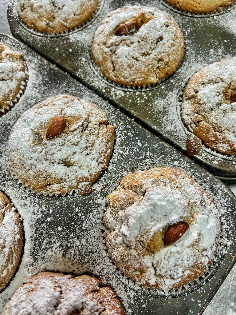 Close-up of muffins in metal baking trays, heavily dusted with powdered sugar. Several muffins have whole almonds on top, with sugar scattered across the tray surfaces.