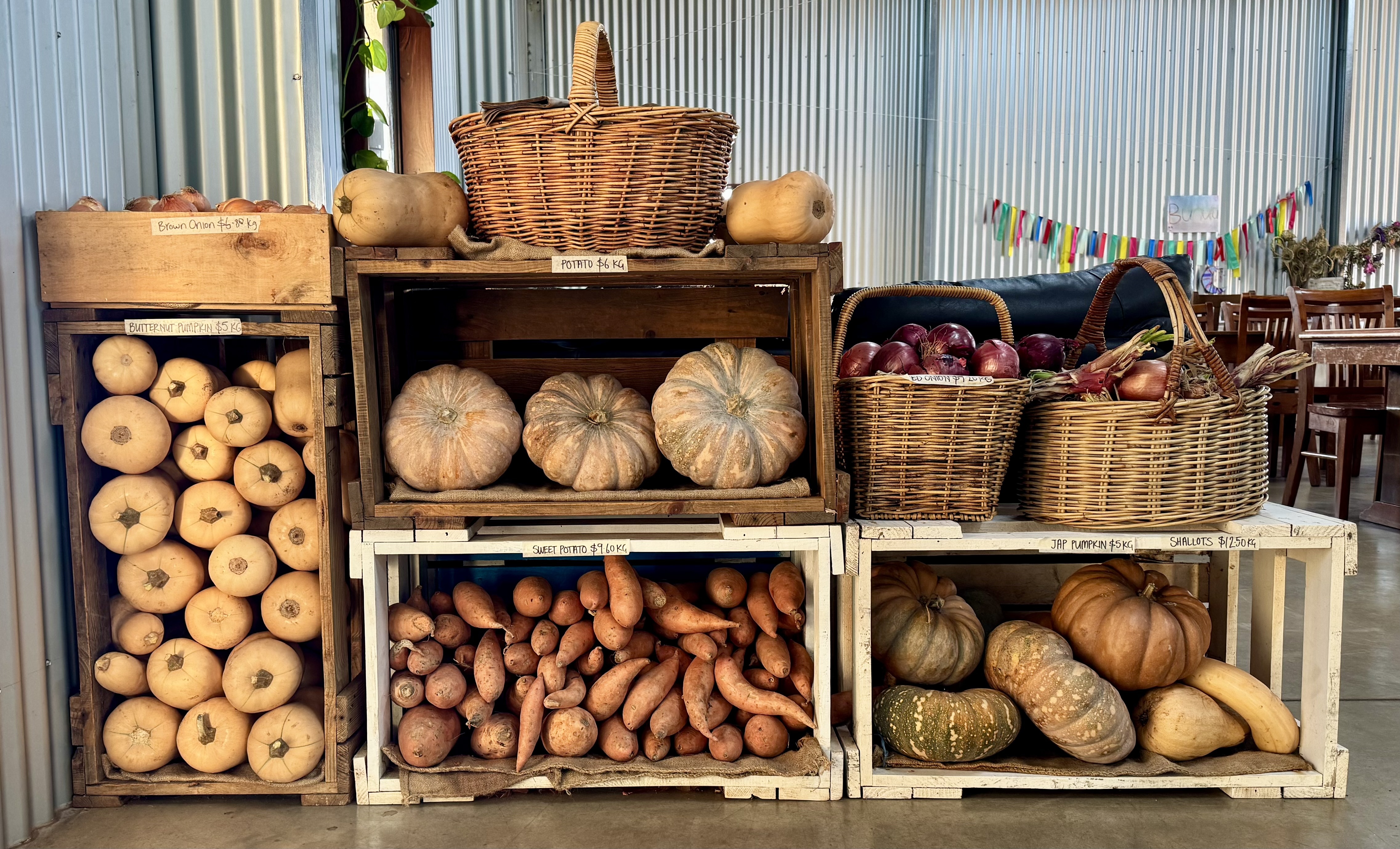 Stacked wooden crates display pumpkins, squash, and sweet potatoes, with wicker baskets of red onions and shallots on top. The produce stand sits against a corrugated metal wall with tables and chairs visible in the background.