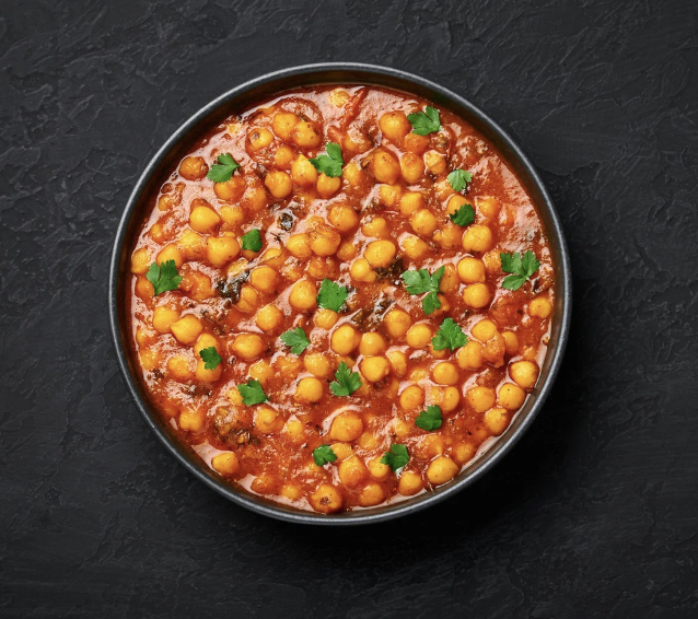 Top-down view of a black bowl filled with chickpeas in a red tomato-based sauce, garnished with small parsley leaves. The bowl sits on a dark textured surface.