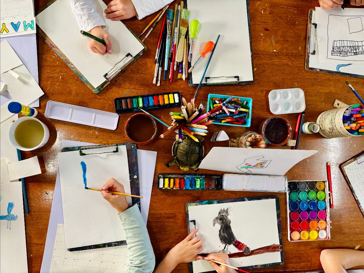 Overhead view of children’s hands drawing and painting on clipboards around a wooden table. The table is scattered with watercolor palettes, brushes, markers, crayons, jars of water, and pencils.