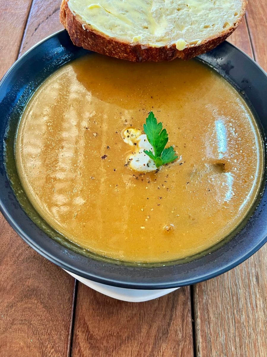 A bowl of creamy brown soup topped with a small dollop of cream, cracked pepper, and a parsley leaf sits on a wooden table. A slice of buttered bread rests on the rim of the bowl.