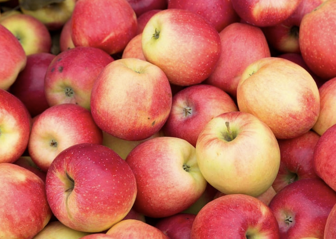 Close-up of a pile of red and yellow apples tightly packed together. Several apples show stems and speckled skin in varying shades of pink, red, and golden yellow.