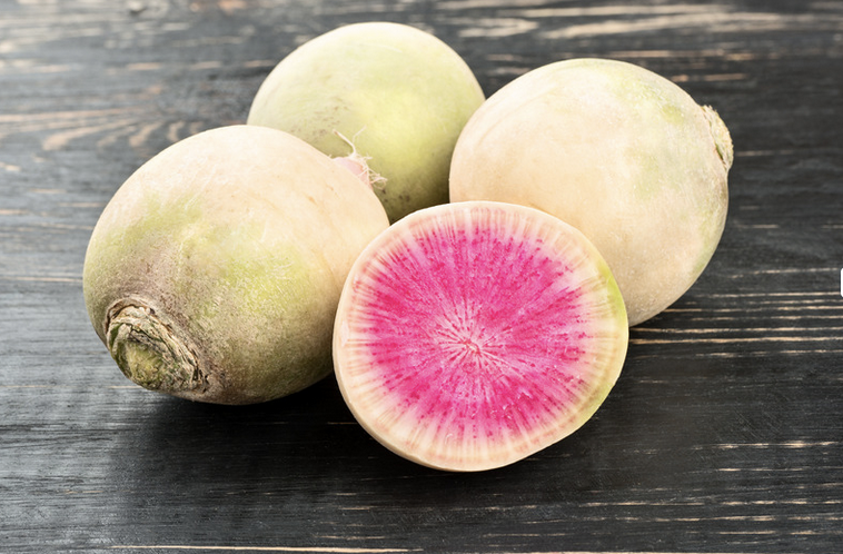 Three pale green-white radishes sit on a dark wooden surface, with one radish cut in half to show a bright pink center and white outer ring.