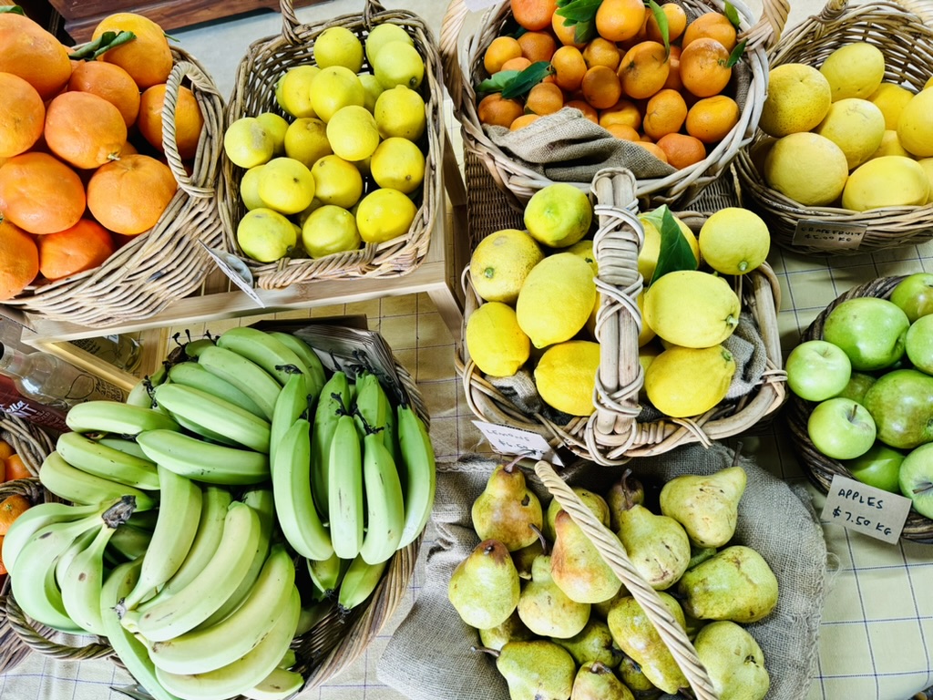 Overhead view of a farm shop display with wicker baskets of citrus fruit (oranges, lemons, and limes), green bananas, pears, and green apples on a tiled counter. Small handwritten price tags are visible beside some baskets.