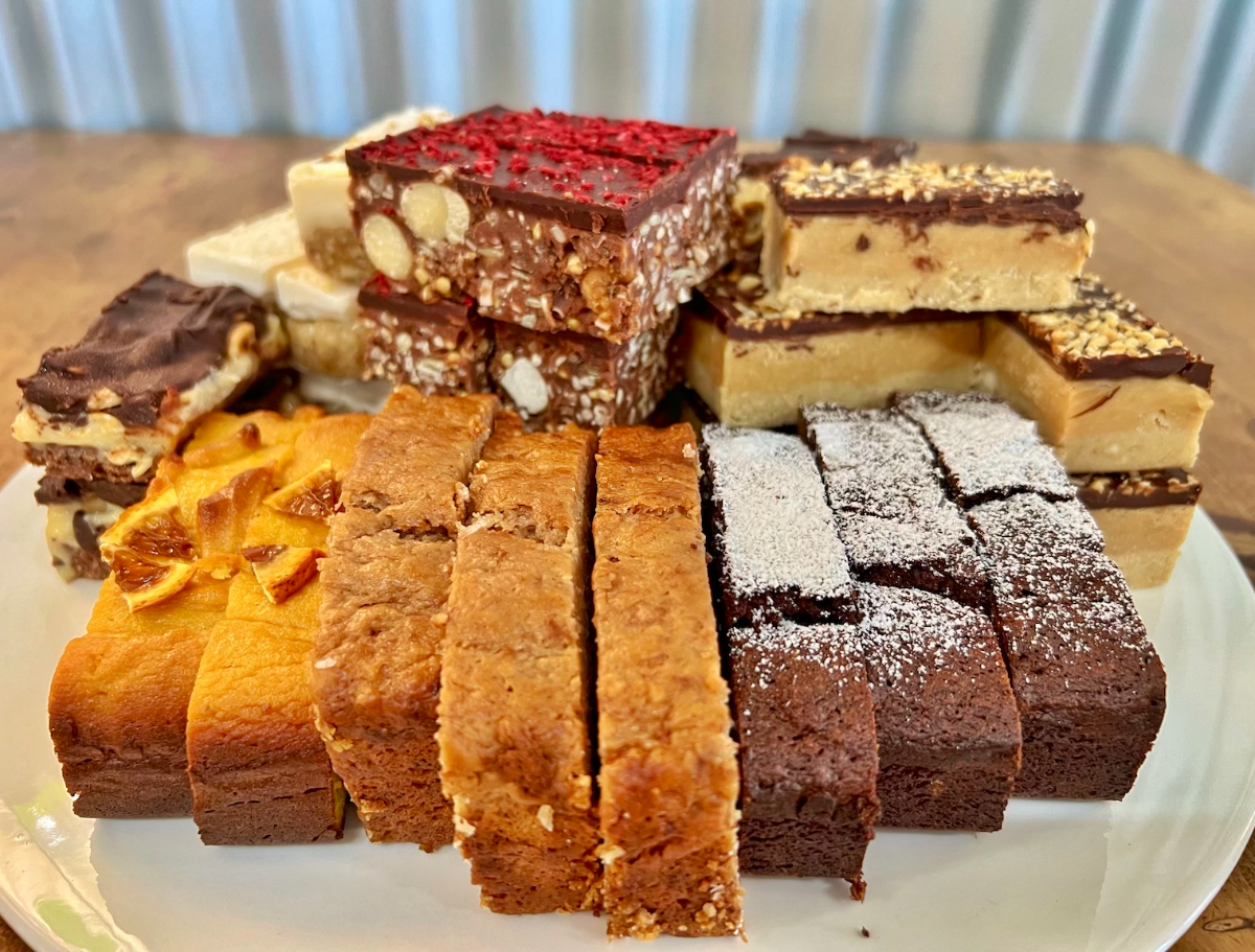 Assorted dessert bars stacked on a white plate, including chocolate-topped nut slices, caramel-colored slices with chopped nuts, and dark brownies dusted with powdered sugar. In front are rectangular blondie-style bars and orange-topped cake slices on a wooden table.